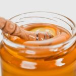 Holzlöffel mit flüssigem Honig über einem Glas mit goldgelber Honigmasse vor weißem Hintergrund