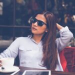Frau mit Sonnenbrille sitzt entspannt im Café mit Kaffee und Tablet, nutzt bewusste Pause zur Regeneration