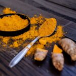 Kurkuma-Pulver in schwarzer Schale und auf Löffel mit frischer Kurkuma-Wurzel auf dunklem Holz