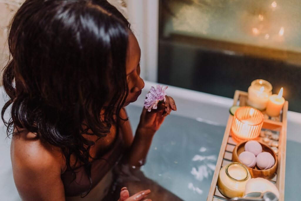 Frau in der Badewanne hält lila Blume, umgeben von Kerzen und Wellness-Produkten für Selbstfürsorge-Routine.