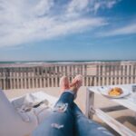 Person entspannt auf Strandterrasse mit Frühstück, Blick auf Meer und Dünen, ideale Wellness-Auszeit zum Abschalten