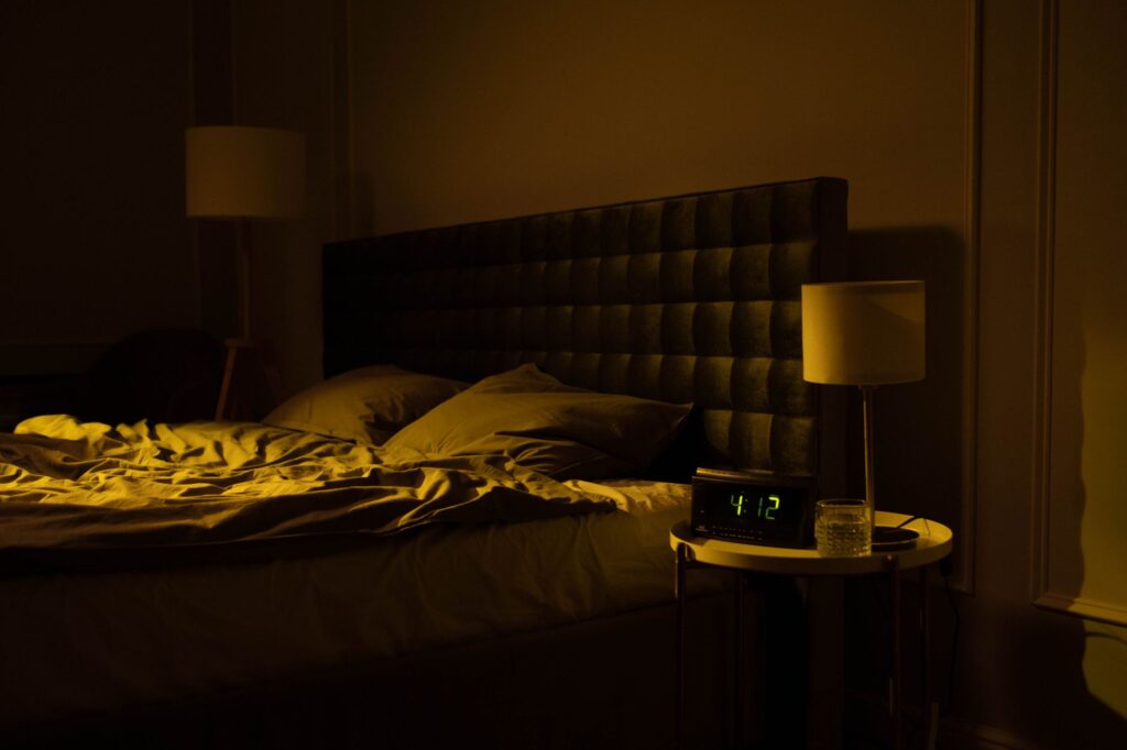 Dunkles Schlafzimmer mit Bett, schwarzem Kopfteil, gelber Nachttischlampe und Wecker zeigt 4:12 Uhr nachts.